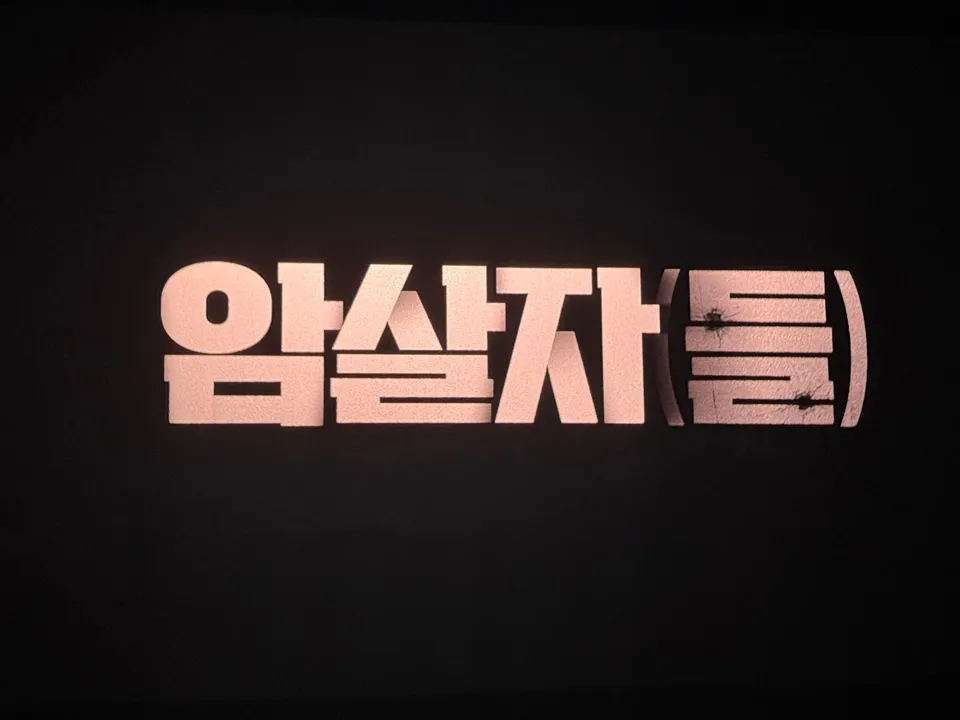 검은 배경에 분홍빛으로 빛나는 '암살자(들)'이라는 글자가 있으며, 괄호 안의 '들' 자에 총구 자국 같은 흔적이 있다.