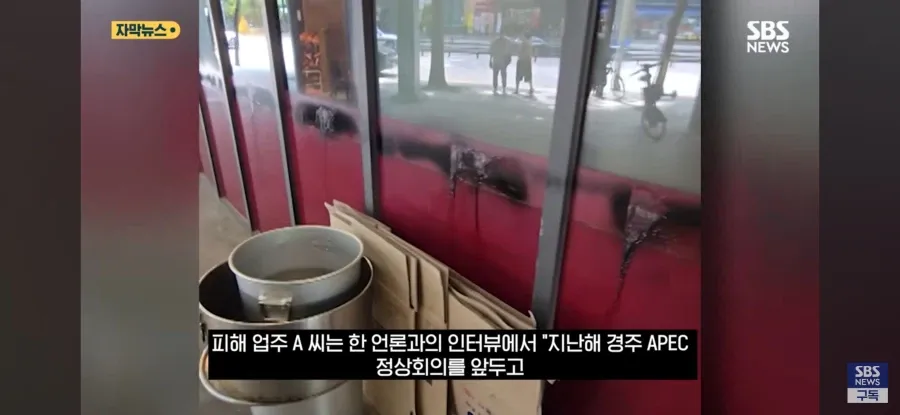 검은색 스프레이로写了字且流淌痕迹的红色店铺门窗，窗前堆放着纸箱和金属锅具， 화면에는 뉴스 자막이 표시되어 있다.